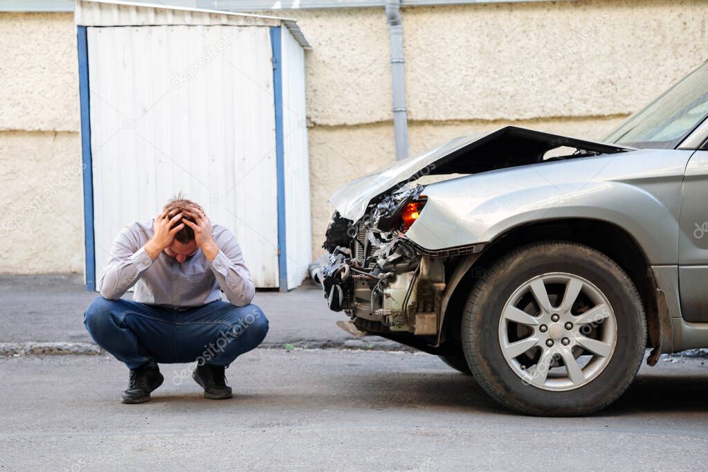 Young man driver in car accident holding his head near broken car on the road after car accident. Caucasian man facepalm holding head injury after accident