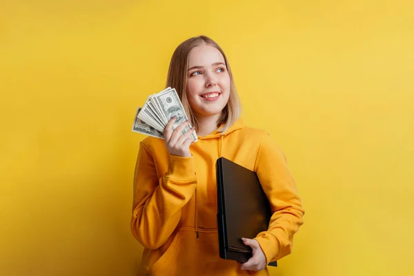 Portrait de jeune jolie femme souriante passionnante tenant une pile de billets d'argent et un ordinateur portable. Adolescente rêvant d'argent, gains en ligne, victoire de loterie isolé sur fond de couleur jaune — Photo