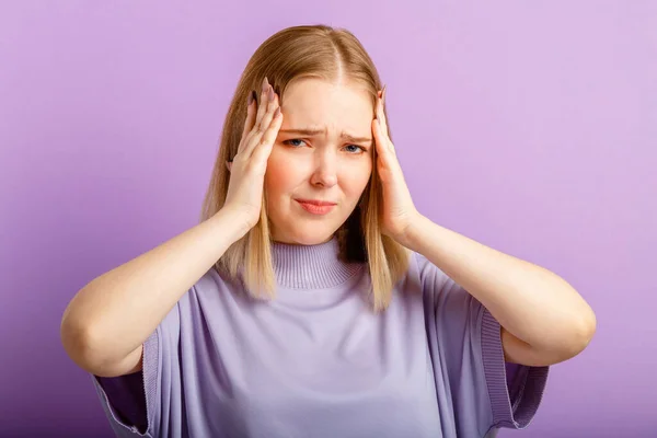Woman experiencing stress headache holding her head. Emotional portrait of sad young woman. Human emotions of pain and regret over isolated color purple background — Stock Photo, Image