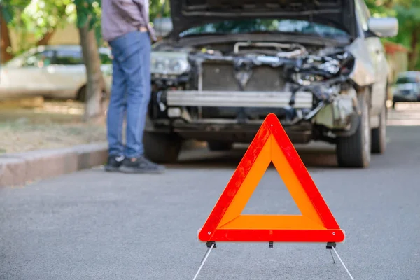 Triangle rouge d'avertissement d'accident de voiture sur la route en face de la voiture naufragée. Un homme inspectant une voiture détruite après un accident. Driver regarder sous le capot de l'automobile naufragée. — Photo