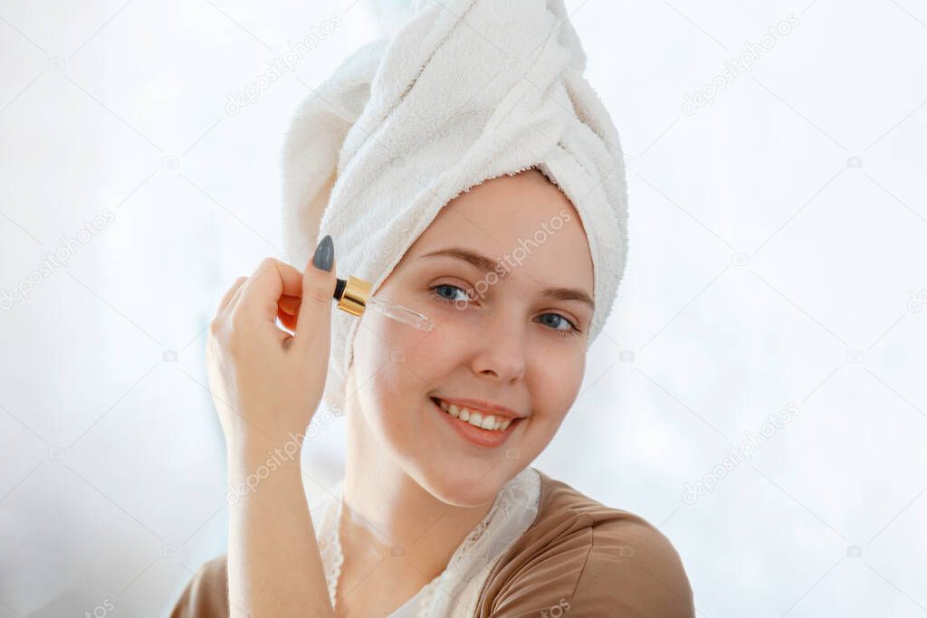 Young beautiful woman portrait applies serum oil in Dropper cosmetic product for health face skin care skin in mirror reflection with towel on head. Self Care as part of morning bathroom routine.
