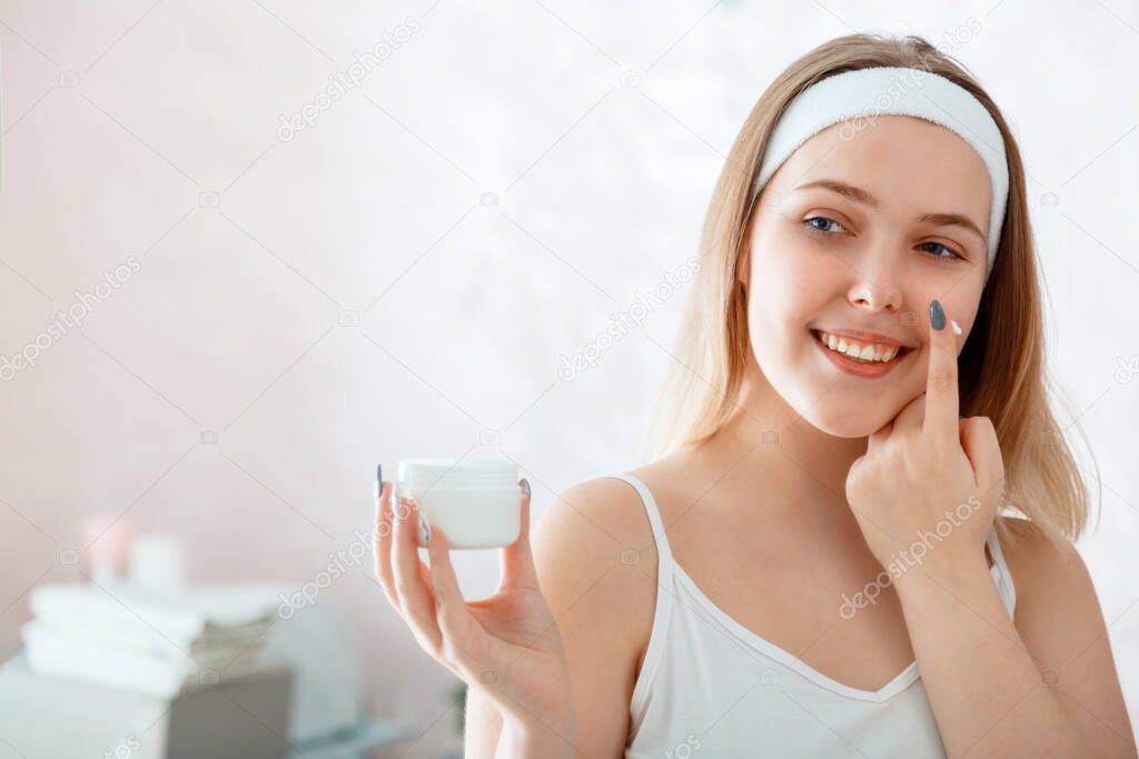 Young beautiful woman holds white jar of moisturizer cream applies cream on face in bathroom. Self Care morning routine. Beauty spa treatment at home. Women use for face skin skincare Beauty products