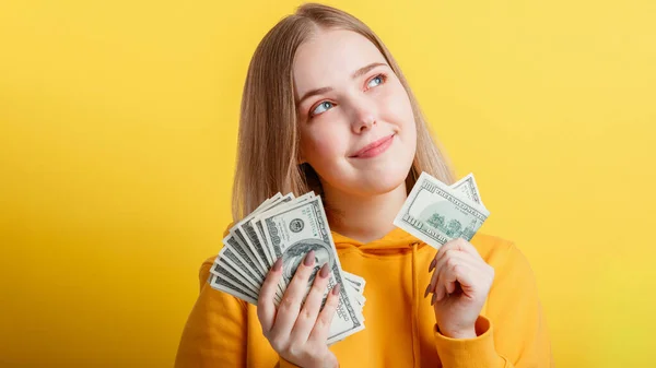 Happy teenage blonde girl holding in hands money cash dollars dreaming thoughtfully isolated on color yellow background. Portrait young excited smiling woman stack of money banknotes. Long web banner — Stock Photo, Image
