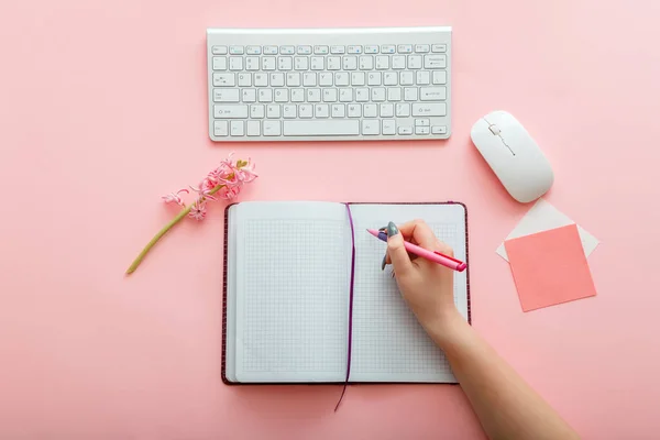 Pink work desk with female hand writes notes in notebook at workplace. Desktop office space top view. Work space girly office table. Back to school layout concept.