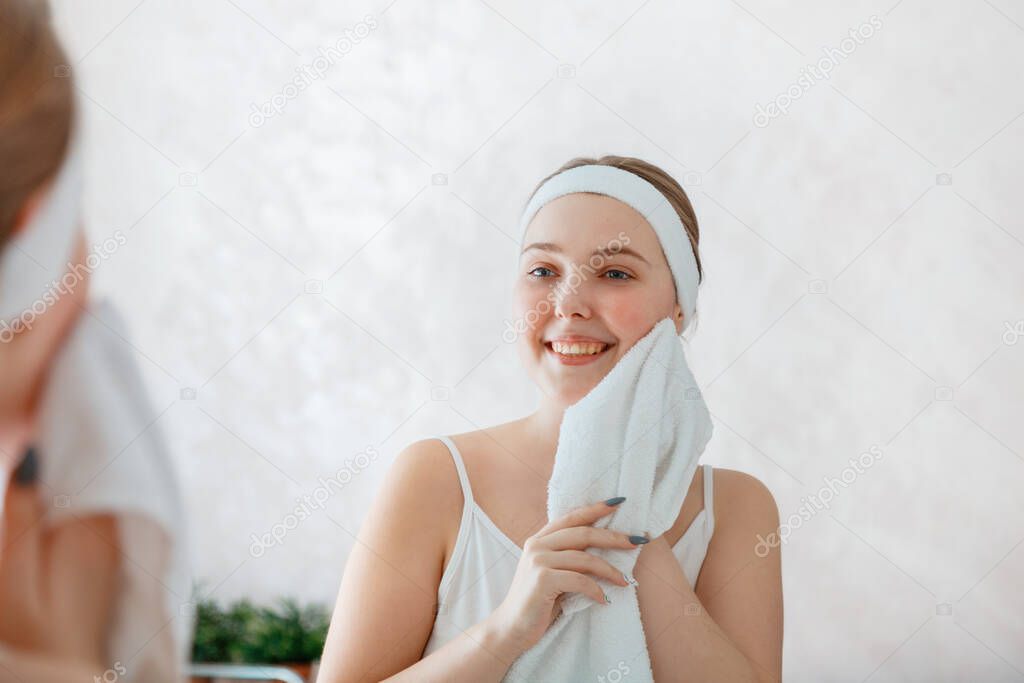 Young woman wipes herself face with white towel after shower washing. Self care in morning at bathroom. Happy smiling teenage girl do morning routine daily hygiene. Portrait reflection in mirror