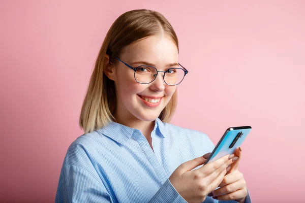 メガネで幸せな正笑顔マネージャーオフィスワーカーの感情的な肖像画は、スマートフォンを使用します。青いシャツの女性はピンクの背景の上に隔離された電話でメッセージを書きます. — ストック写真