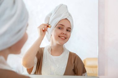 Genç ve güzel bir kadın aynanın yanındaki banyoda cilt bakımı için damlatıcıya yağ serumu sürüyor. Sabah rutininde kişisel bakım kozmetik ürünü. Kafasında havlu olan genç ve güzel bir kız..