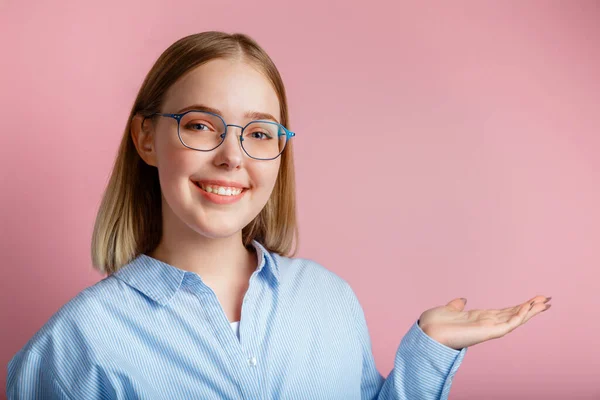 Ung affärskvinna eller chef i glasögon kontorskläder ler visa sida gest för hand. Tonåring flicka student bär glasögon visar hand gest på kopia utrymme isolerad över färg rosa bakgrund — Stockfoto