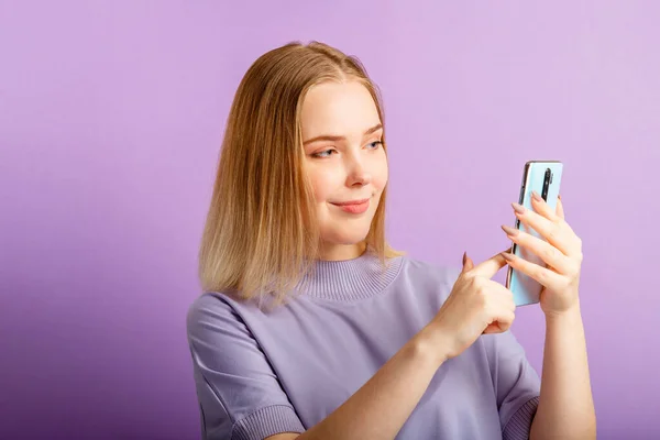 若い女性は、孤立した色の紫色の背景に携帯電話を保持アプリを介してメッセージを送信スマートフォンを使用します. — ストック写真