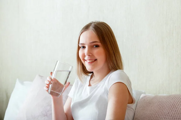 Smile young woman drinks glass of pure water in morning after waking up in her bed room. Happy teen girl maintains water balance for body health by drinking clean water.