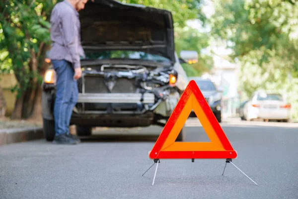 Autóbaleset piros háromszög figyelmeztető jele. Háromszög az összetört autó és a sofőr előtt. Sérült ember baleset után belenéz a motorháztető alá.. — Stock Fotó