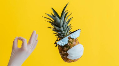 Eldivenli el, tamam işareti gösteriyor. Tıbbi yüz maskeli ananas, sarı arka planda güneş gözlüğü. Tecrit süresince seyahat konseptinde yaz arkaplanındaki ananas meyvesi. Uzun web pankartı
