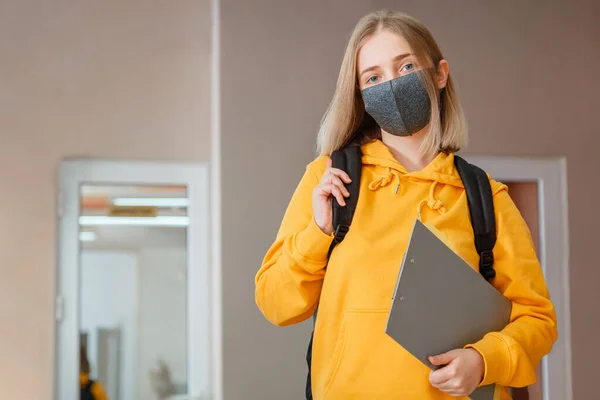 Estudante em máscara com mochila e bloco de notas. Jovem estudante em máscara médica protetora. Retrato de estudante loira Menina no interior da universidade durante coronavírus covid 19 lockdow. — Fotografia de Stock