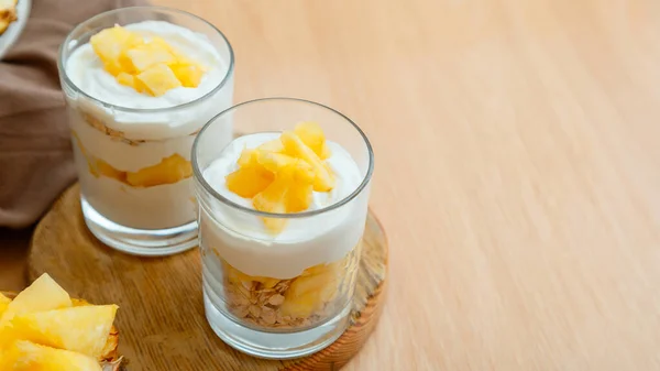 Tasty pineapple desserts with chopped fresh juicy pineapple. Breakfast dessert with oat granola, greek yogurt and pineapple in layers in glass on wooden table. Long web banner with copy space.