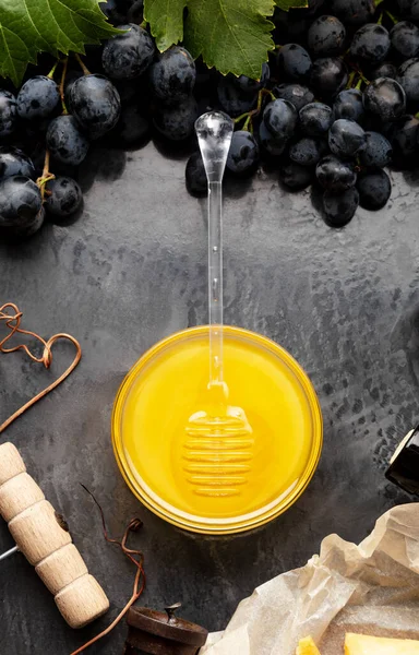 Honey in glass jar with honey spoon on dark stone board with wine cheese corkscrew in Black juicy grapes frame. Delicious yellow bee honey in snack plate on black table. Top view. Vertical photo — Stock Photo, Image