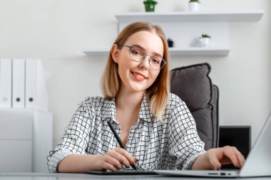 Genç gülümseyen kadın ofis içi bilgisayarını kullanarak grafik tablete resim çiziyor. Kafkas Sarışın genç mutlu kadın gözlüklü serbest çizer Grafik Tasarımcısı.