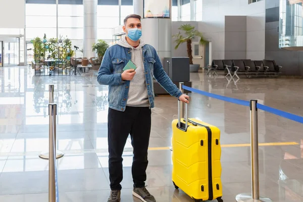 Homem em máscara facial médica na frente do balcão de check-in no interior do aeroporto durante o bloqueio vívido. Viajante casual urbano detém bagagem de mala de passaporte antes de embarcar em um voo aéreo. — Fotografia de Stock