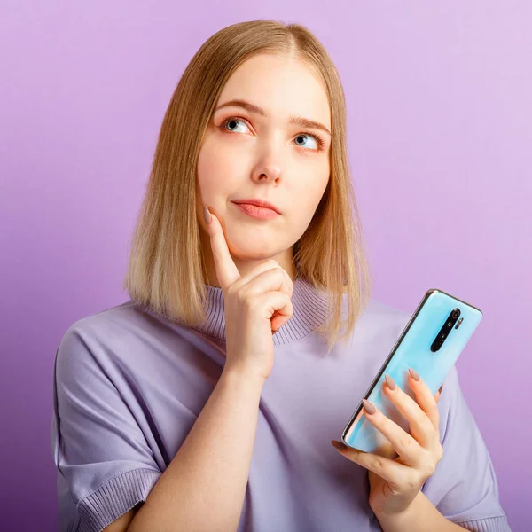 Young woman think doubt about question using smartphone. Teen girl curious dreaming, choose or decide look side on copy space. Emotion portrait isolated over purple color background. Closeup square — Stock Photo, Image