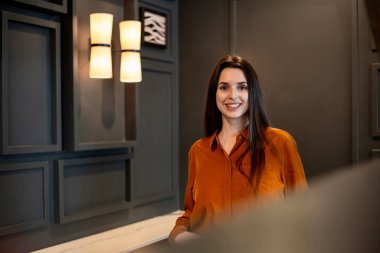 Karanlık bir duvarla modern bir iç mekanda oturmuş gülümseyen bir kadın, rahat ve dostane bir ifadeyle rahatça poz veriyor, sıcak ve doğal bir portre oluşturuyor..