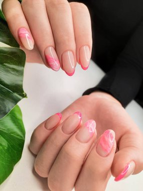 Soyut tasarımlı güzel pembe Fransız manikürü. Yeşil yapraklı arka planda güzel uzun tırnakları olan kadın parmakları.