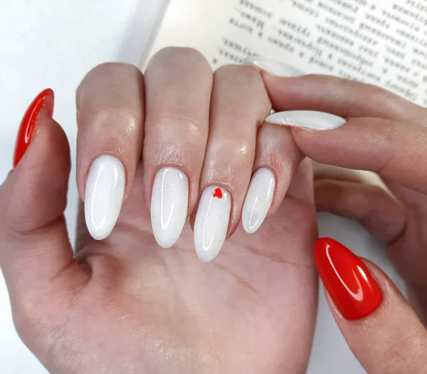 White nails with a red heart design on the background of the book text. Women\'s hands with a white manicure with a red nail and a heart.