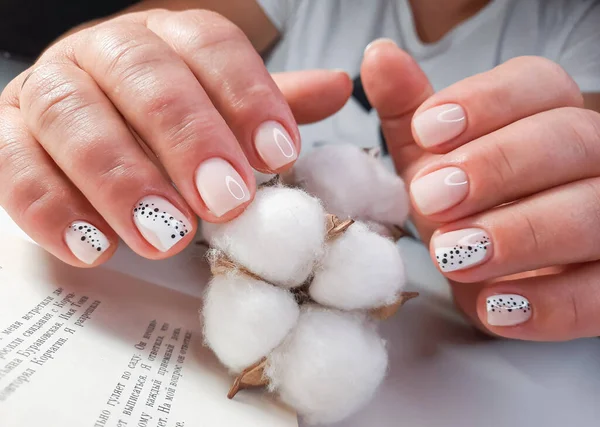 Soft pink gel polish with a design of black dots on the background of the text of the book. Women\'s hands with pastel colored nails hold a sprig of cotton. Milk manicure with black and white design.