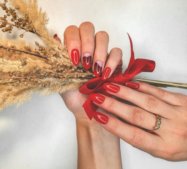 Red nails with a burgundy gradient on the background of a yellow twig with a red bow. Beautiful manicure with gel polish coating with a dry branch in your hand