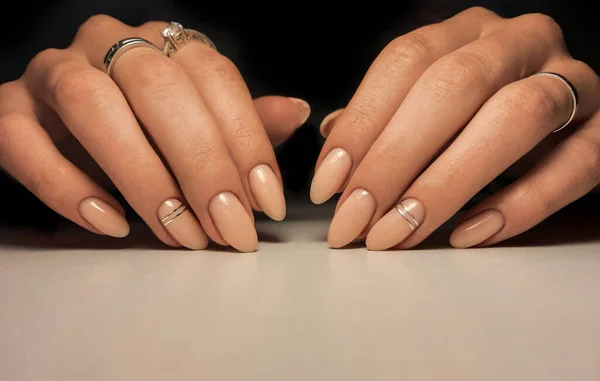 Gentle gel polish of beige color on almond-shaped nails with silver stripes. Nude manicure with shiny stripes design. Pastel color coating on the nails.
