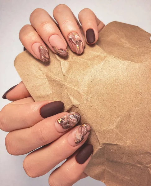 A charming chocolate gel polish with a fluid abstract design. Women\'s hands with camouflage-colored manicure and brown design hold a kraft bag. Pastel color coating on the nails with brown marble.