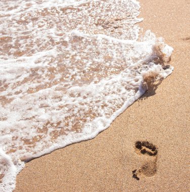 Kumda deniz suyuyla insan ayak izleri. Kumun üzerindeki bir dalgadan köpük. Köpüklü bir okyanus dalgası deniz kabuğu sahilindeki izleri temizler.