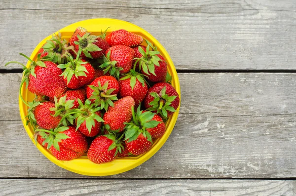 Summer berries topic: Ripe red strawberries lying in a yellow plate on a pile of gray wooden table in the garden, view from above