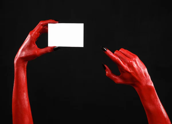 Tema de Halloween: Mão do diabo vermelho com pregos pretos segurando um cartão branco em branco em um fundo preto — Fotografia de Stock