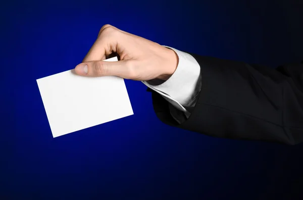 Tema comercial y publicitario: Hombre de traje negro sosteniendo una tarjeta blanca en blanco en su mano sobre un fondo azul oscuro en estudio aislado — Foto de Stock