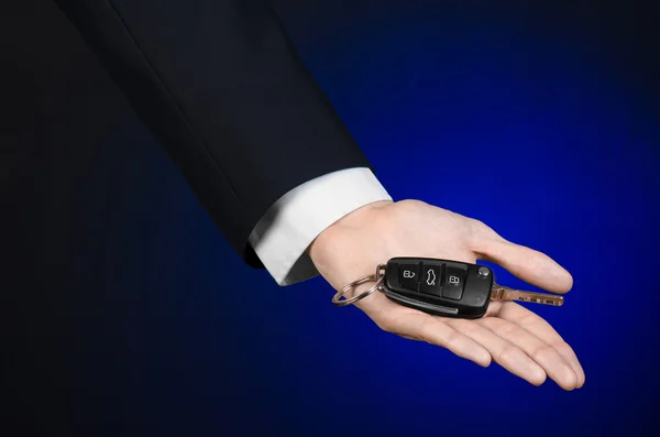 Business and gift theme: car salesman in a black suit holds the keys to a new car on a dark blue background in studio — Stock Photo, Image