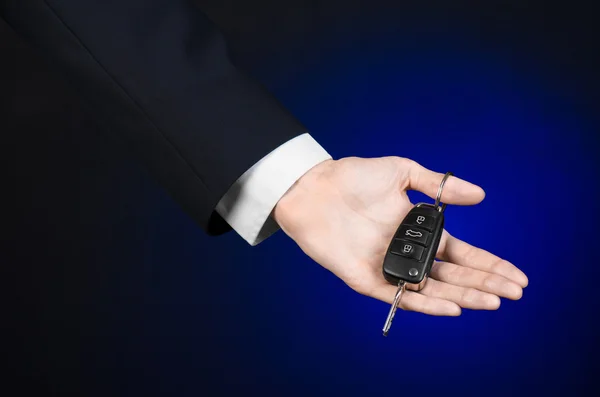 Business and gift theme: car salesman in a black suit holds the keys to a new car on a dark blue background in studio — Stock Photo, Image