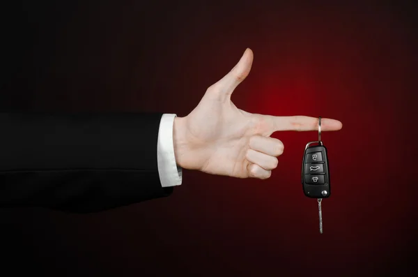 Business and gift theme: car salesman in a black suit holds the keys to a new car on a dark red background in studio — Stock Photo, Image
