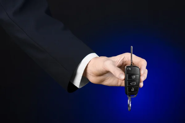 Business and gift theme: car salesman in a black suit holds the keys to a new car on a dark blue background in studio — Stock Photo, Image