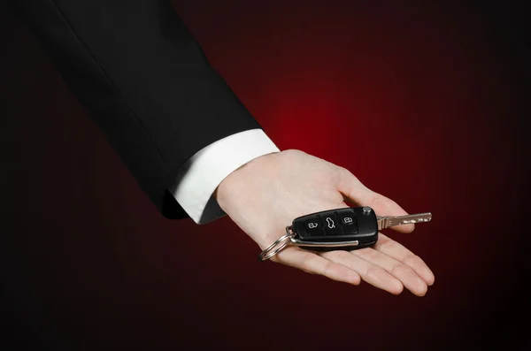 Tema de negocios y regalos: vendedor de coches en un traje negro sostiene las llaves de un coche nuevo sobre un fondo rojo oscuro en el estudio —  Fotos de Stock