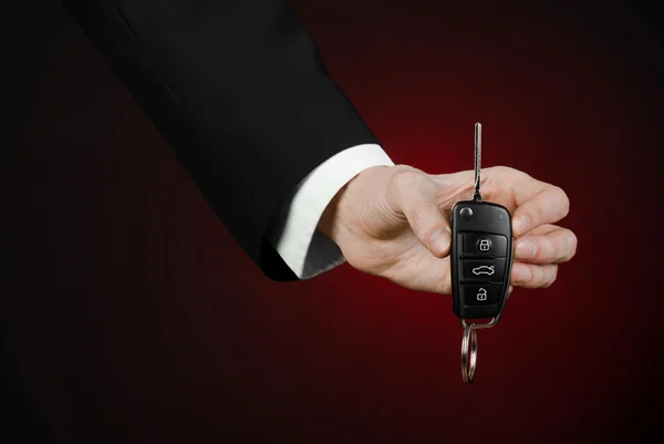 Business and gift theme: car salesman in a black suit holds the keys to a new car on a dark red background in studio — Stock Photo, Image