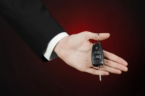 Business and gift theme: car salesman in a black suit holds the keys to a new car on a dark red background in studio — Stock Photo, Image