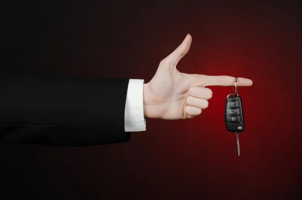 Tema de negocios y regalos: vendedor de coches en un traje negro sostiene las llaves de un coche nuevo sobre un fondo rojo oscuro en el estudio —  Fotos de Stock