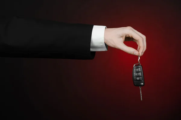 Business and gift theme: car salesman in a black suit holds the keys to a new car on a dark red background in studio — Stock Photo, Image