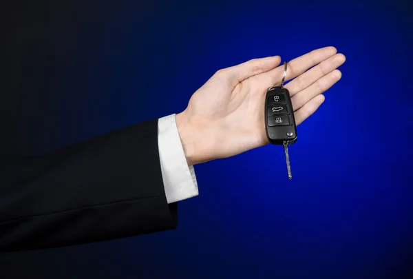 Tema de negocios y regalos: vendedor de coches en un traje negro sostiene las llaves de un coche nuevo sobre un fondo azul oscuro en el estudio —  Fotos de Stock