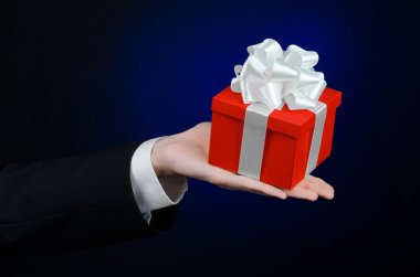 Kutlamalar ve hediye Tema: Studio izole koyu mavi zemin üzerine beyaz kurdele, güzel kırmızı kutu sarılı özel hediye ve pahalı hediye tutarak siyah takım elbiseli bir adam