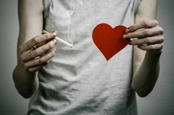 Cigarettes, addiction and public health topic: smoker holds the cigarette in his hand and a red heart on a dark background in the studio — Stock Photo, Image