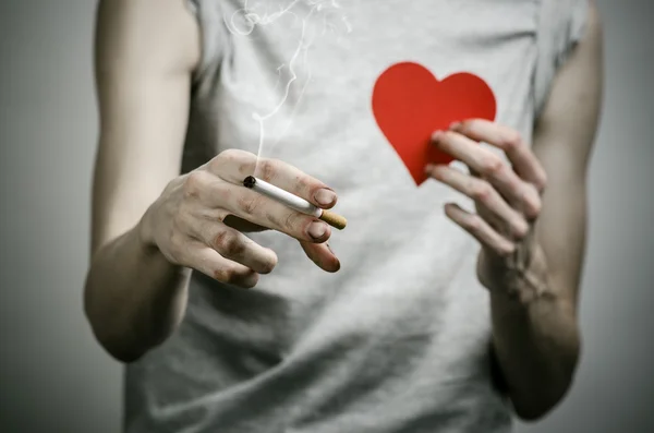Zigaretten, Sucht und öffentliches Gesundheitsthema: Raucher hält Zigarette in der Hand und rotes Herz auf dunklem Hintergrund im Studio — Stockfoto