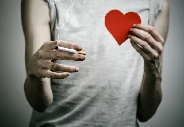 Cigarettes, addiction and public health topic: smoker holds the cigarette in his hand and a red heart on a dark background in the studio — Stock Photo, Image