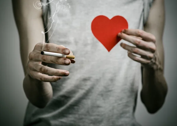 Cigarettes, addiction and public health topic: smoker holds the cigarette in his hand and a red heart on a dark background in the studio — Stock Photo, Image