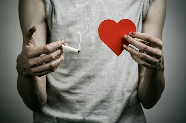 Cigarettes, addiction and public health topic: smoker holds the cigarette in his hand and a red heart on a dark background in the studio — Stock Photo, Image