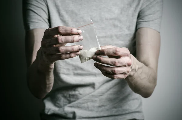 A luta contra as drogas e o tema da toxicodependência: viciado segurando pacote de cocaína em uma camiseta cinza em um fundo escuro no estúdio — Fotografia de Stock